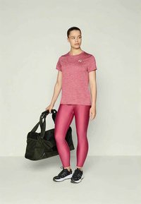 Kvinne i rosa treningsgenser og blanke leggings holder en svart treningsveske, iført svart og hvite joggesko, står foran en ensfarget bakgrunn.