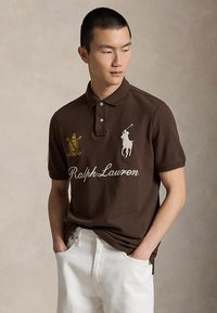 Νέος άντρας που φορά καφέ polo Ralph Lauren με κεντημένα λογότυπα, λευκό παντελόνι, χέρια στις τσέπες, κοιτάζοντας αριστερά πάνω σε απλό φόντο.