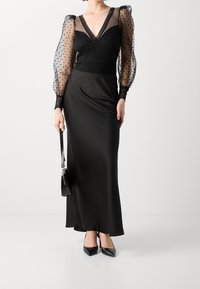 Donna che indossa un lungo abito nero con corsetto in pizzo, maniche a sbuffo trasparenti con pois, tacchi a punta neri e tiene in mano una piccola borsa nera.