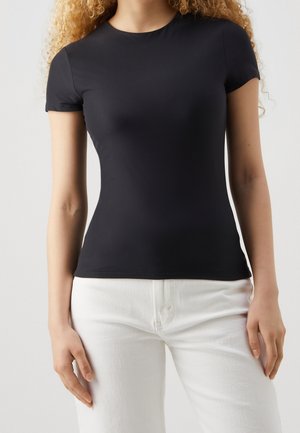 Nainen, jolla on päällään istuva musta lyhythihainen paita ja korkea vyötäröiset valkoiset housut, seisoo yksinkertaisen vaalean taustan edessä.