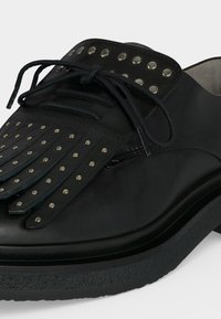Chaussure de ville en cuir noir avec un panneau décoratif à franges et des clous argentés, équipée de lacets noirs et d'une semelle épaisse texturée.