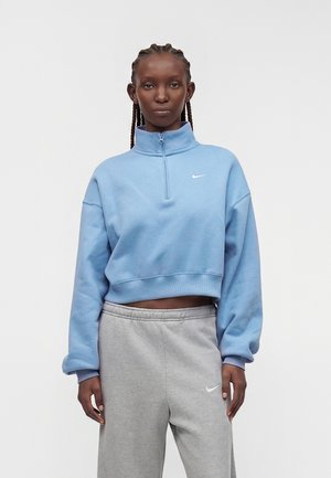 Femme avec des cheveux tressés portant un sweat-shirt bleu clair à fermeture éclair quart de zip et un pantalon de jogging gris, debout sur un fond blanc uni.