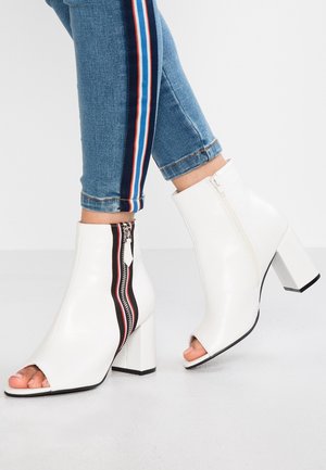 Białe botki na kostkę z sztucznej skóry z odkrytymi palcami i bocznym zamkiem, z czarnymi i czerwonymi akcentami. Kwadratowy obcas, gładka tekstura, smukły design.