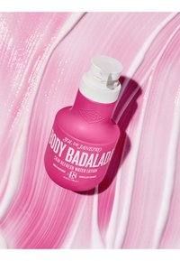 Botella de color rosa brillante de loción Sol de Janeiro Body Badalada con bomba blanca, colocada sobre un fondo de textura cremosa en tonos rosa y blanco.