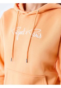 Oranje hoodie met een trekkoord, voorzien van een witte geborduurde "Project x Paris" logo op de voorkant. Zacht materiaal en een buidelzak.