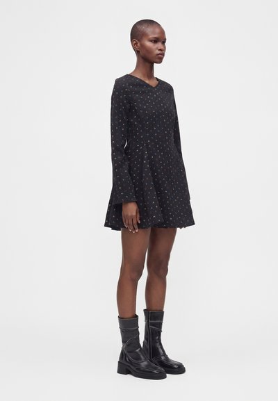 Femme portant une robe noire à manches longues avec de petits pois colorés et des bottes en cuir noires mi-mollet, debout devant un fond uni.