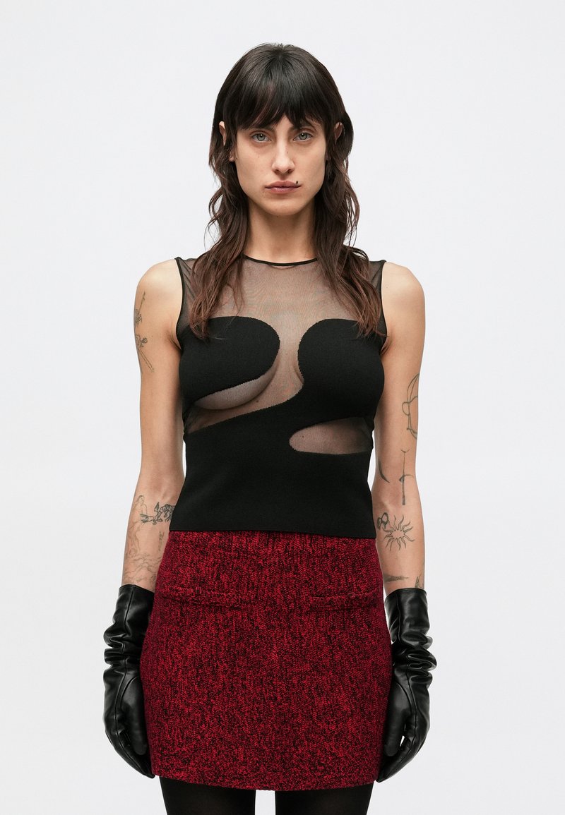Czarny bezrękawnik z przezroczystymi panelami ozdobiony wycięciami. Czerwona spódnica o fakturze i czarne skórzane rękawiczki, minimalna ilość dodatków.