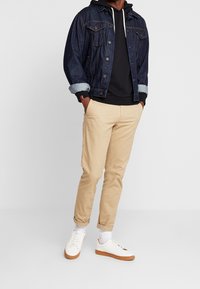 Jeansjacka med mörk tvätt och uppvikta ärmar, tillsammans med en svart hoodie, beige byxor och vita sneakers med bruna sulor.