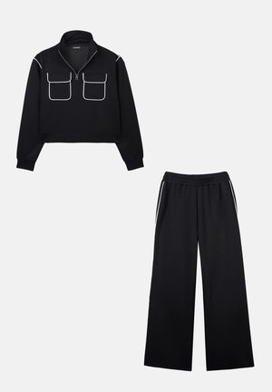 Pullover nero corto con collo a zip e due tasche frontali; abbinato a pantaloni neri a gamba larga con vita elasticizzata e cordoncino.
