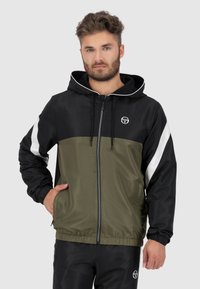 Schwarze und olivgrüne Windjacke mit Reißverschluss, Kapuze und verstellbaren Kordeln. Mit weißen seitlichen Akzenten und elastischen Bündchen.