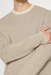 Beige a černý pruhovaný pletený svetr na osobě se zkříženýma rukama, zkrácený nad ústy a pod pasem.