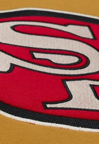 Rotes und schwarzes besticktes "S" auf einem tanfarbenen Stoffhintergrund mit glatter Textur und kontrastierender Naht.