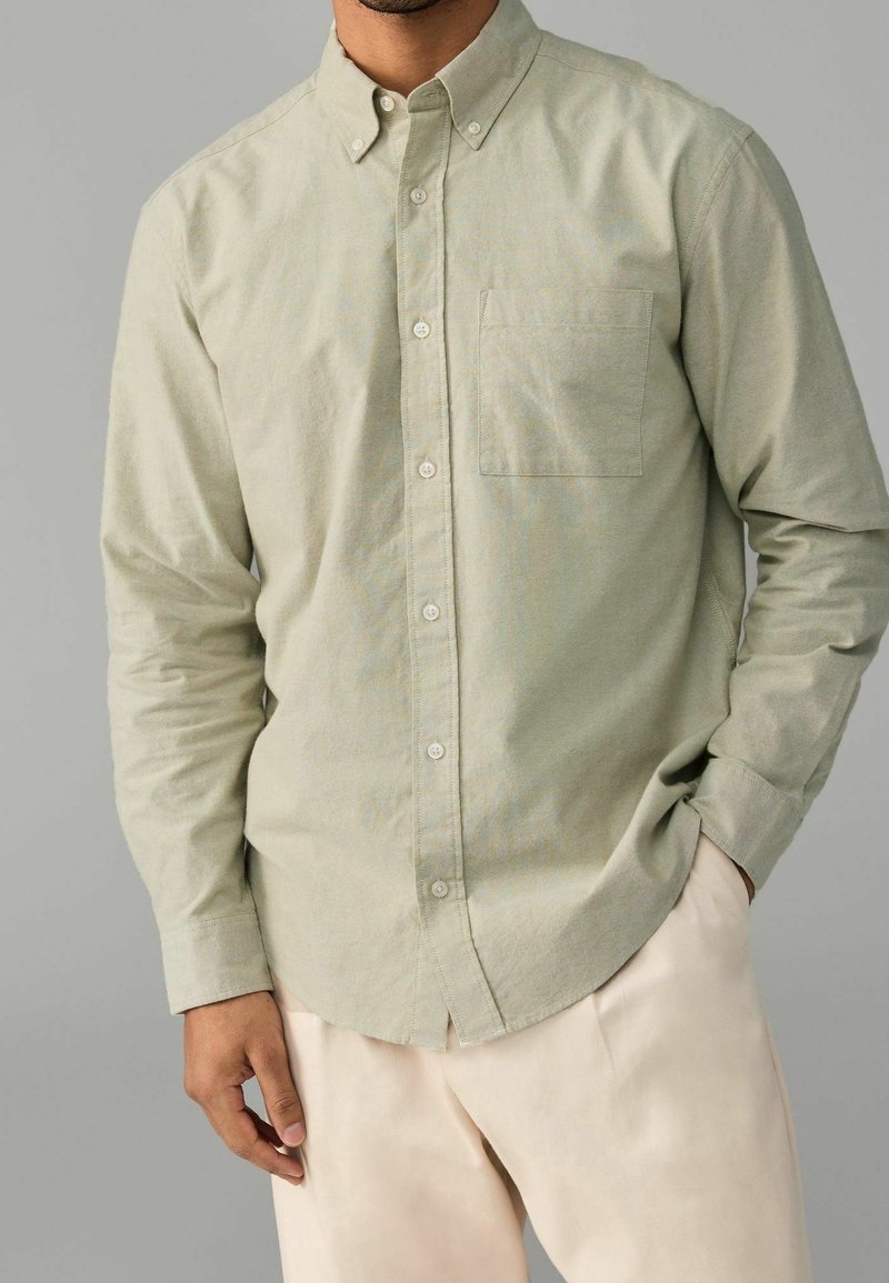 Homme portant une chemise beige clair à manches longues boutonnée avec une poche poitrine et un pantalon clair, main dans la poche sur fond gris.
