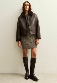 Giacca cropped in pelle marrone con colletto in pelliccia, abbinata a un vestito a maglia beige testurizzato e stivali in pelle marrone al ginocchio.