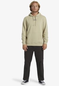 Sudadera beige clara con un bolsillo tipo canguro y capucha con cordón, combinada con pantalones oscuros y zapatos en blanco y negro. Diseño sencillo, sin patrones.