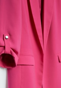 Blazer rosa brillante con las mangas remangadas aseguradas por una pestaña de botón y bolsillos de solapa visibles en la parte frontal.