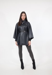 Schwarzer Poncho aus Kunstleder mit Kragen, an der Taille mit einem Gürtel geschnürt, getragen über grauen Skinny-Jeans und schwarzen Stiefeletten.