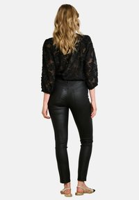 Zwarte doorzichtige blouse met een textuurrijk bloemenpatroon, strakke zwarte lerenachtige broek en tan sandalen. Lang golvend haar zichtbaar van achteren.