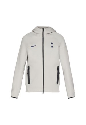 Svijetlosiva dukserica na zip, s Nike logotipom i Tottenham emblemom, s crnim patentnim zatvaračem i detaljima, glatkog materijala i dizajnom s kapuljačom.