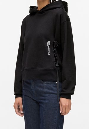 Osoba ubrana w czarną bluzę z kapturem z koronkowym detalem po bokach oraz białym napisem „Karl Lagerfeld Jeans” w połączeniu z ciemnoniebieskimi dżinsami na białym tle.
