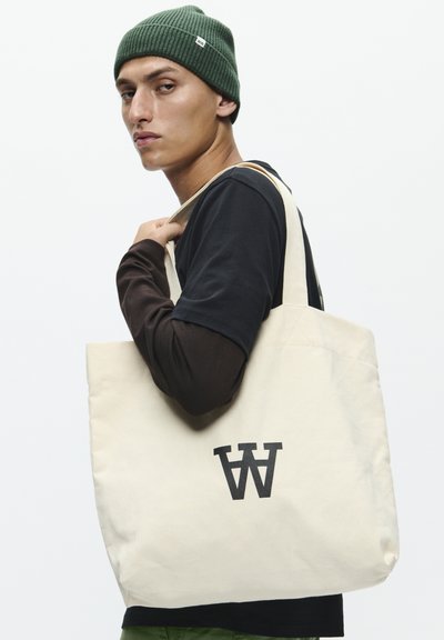 Sac fourre-tout en toile beige avec un audacieux logo "W" noir, doté de deux poignées robustes. Le modèle porte une chemise noire et un pantalon vert.