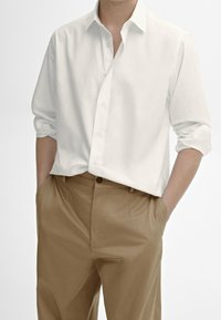 Chemise blanche à boutons avec manches retroussées, associée à un pantalon sur mesure marron clair, tous deux présentant des textures lisses et des lignes épurées.