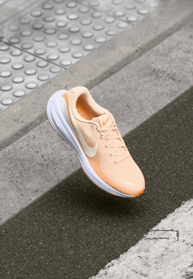 Chaussure de course Nike orange clair avec semelle blanche et logo swoosh, positionnée au-dessus d’un revêtement urbain à surface texturée et ligne blanche peinte.