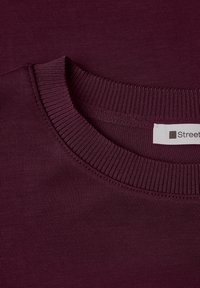 Gros plan d'un col rond côtelé sur un sweat-shirt violet foncé avec une étiquette blanche de vêtement partiellement visible.