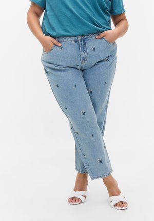 Person trägt hellblaue Jeans mit kleiner Blumenstickerei und weiße offene Sandalen mit geflochtenen Riemen, steht mit verschränkten Beinen und hat die Hände in den Taschen.