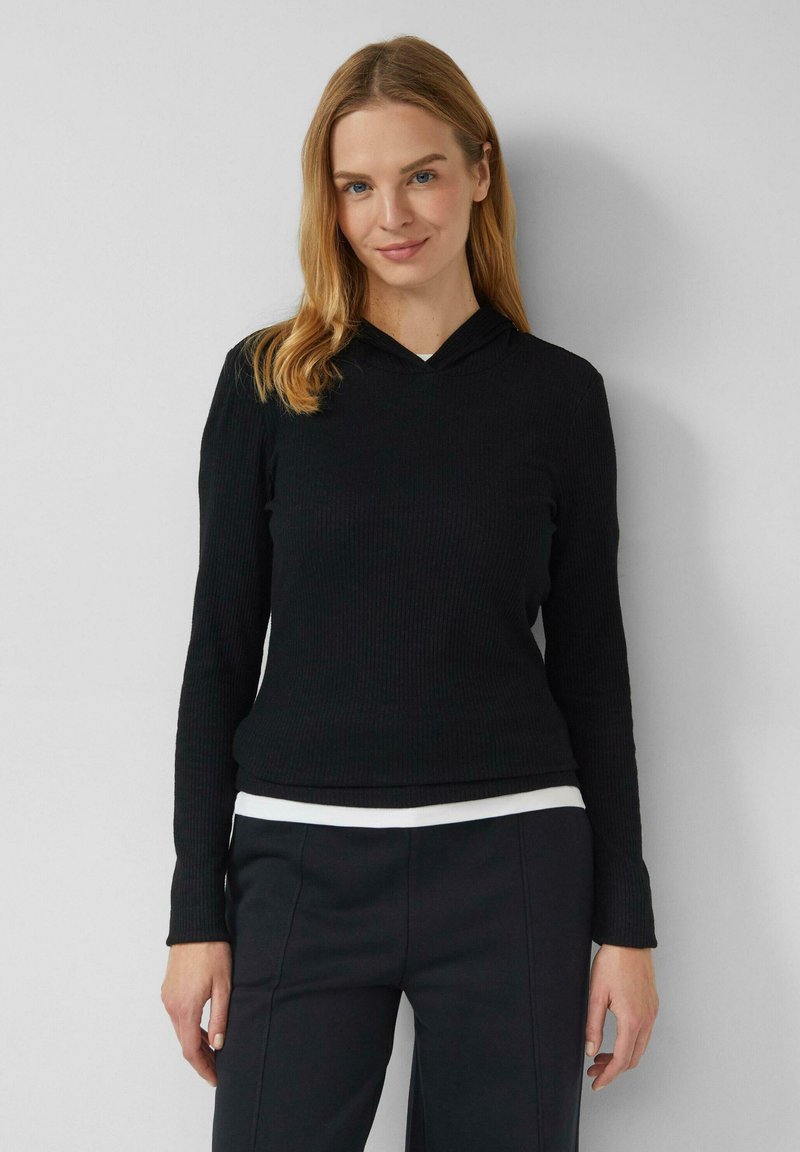 Czarna prążkowana bluza z długim rękawem i kapturem, z dekoltem w kształcie litery V oraz miękką fakturą, noszona z białą koszulą widoczną poniżej. Stylizację uzupełniają czarne spodnie.