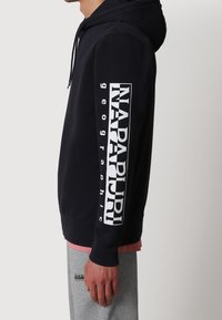 Persona che indossa un hoodie nero con la scritta "NAPAPIJRI geographic" sulla manica e pantaloni felpati grigi con il logo vicino alla tasca, in piedi di lato.