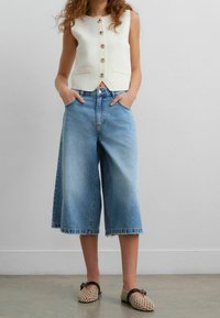 Chaleco sin mangas blanco con botones dorados, combinado con culottes de mezclilla anchos en azul claro y zapatos planos de rafia beige con acentos negros.