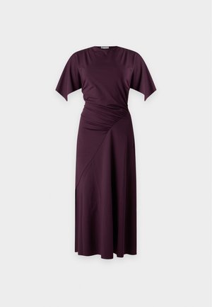 Donna con lunghi capelli neri che indossa un vestito midi arricciato di colore viola scuro e tacchi neri con cinturino alla caviglia, in piedi davanti a uno sfondo semplice.