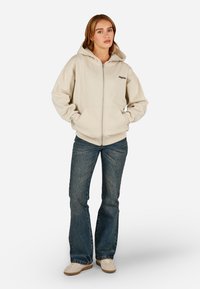 Sweatshirt com fecho em bege e capuz largo, apresentando um logótipo no lado esquerdo do peito. Usado com jeans azuis de perna larga e sapatos de tonalidade clara.