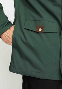 Groene jas met een gladde textuur, voorzien van een borstzak met een bruine klep en een metalen drukknoop. De zoom is zichtbaar aan de onderrand.