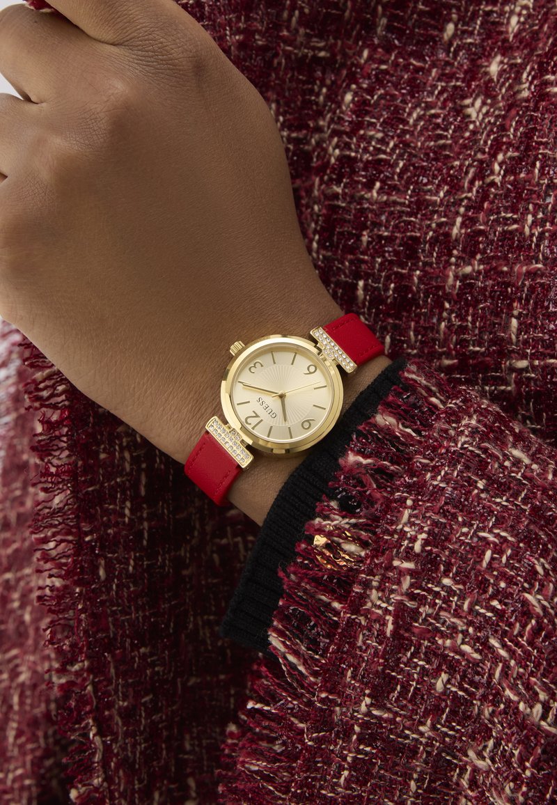 Gold-tone watch with a round face, red leather strap, and rhinestone accents. Texture features a ribbed yellow-gold dial. Worn on a wrist.