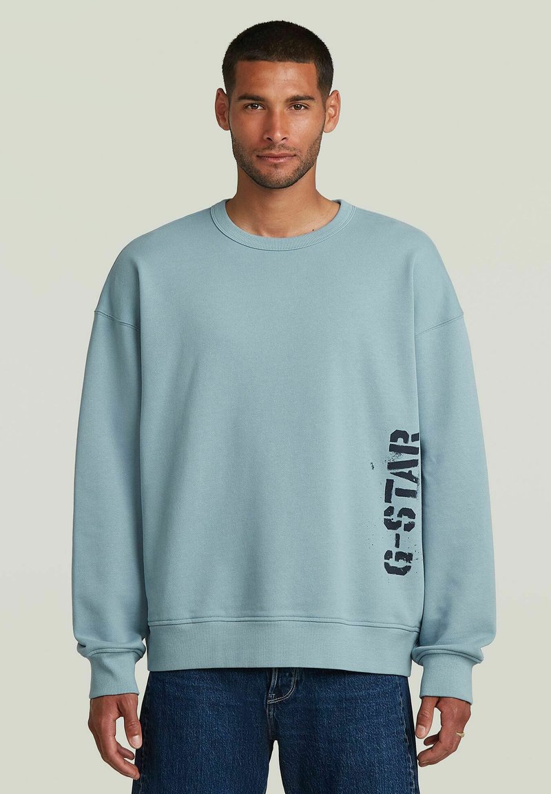 Sudadera azul clara con cuello redondo, hombros caídos y un logo negro en la parte inferior derecha. Textura suave, puños acanalados.