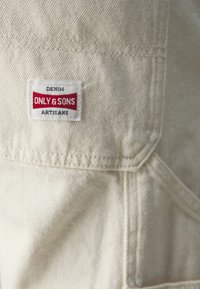 Denim tkanina boje kreme s vidljivom teksturom, koja sadrži ušivenu oznaku s natpisom "DENIM ONLY & SONS ARTISANS" u crvenoj i plavoj boji.