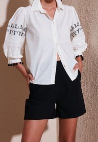 Chemise blanche à manches bouffantes, ornée de broderies florales noires et d'accents en pompons. Assortie à un short noir. Tissu texturé.