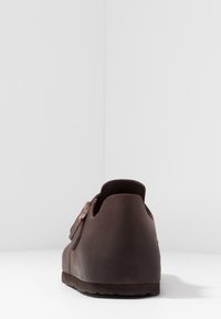 Brown leather shoe with a rounded shape, featuring a strap and buckle detail. The sole is textured, providing grip.