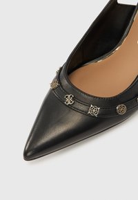 Pantof negru din piele cu vârf ascuțit, cu accente metalice argintii, inclusiv web-uri și un logo, cu o textură netedă și părți laterale deschise.