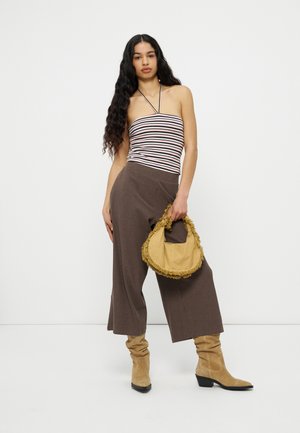 Femme aux longs cheveux bouclés portant un haut rayé à col licou, un pantalon large marron, des bottes marron clair décontractées, tenant un sac à main jaune à franges sur fond blanc.