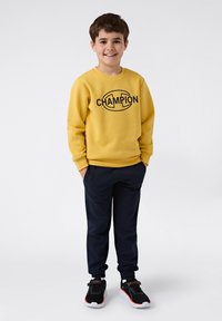 Geel sweatshirt met het zwarte "CHAMPION" logo, gecombineerd met marineblauwe trainingsbroek en zwarte sneakers met rode accenten. Jongentje met een glimlach.