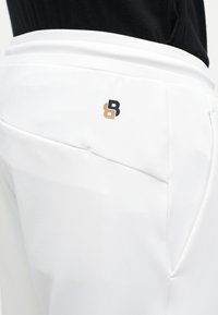 Κοντινό πλάνο άσπρων παντελονιών με μικρό μπεζ και μαύρο λογότυπο κοντά στην πίσω τσέπη, φορεμένα με μαύρο τοπ.