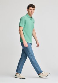 Camisa polo verde de manga corta, con tapeta de botones, combinada con jeans de mezclilla azul claro y zapatillas beige con acentos verdes.