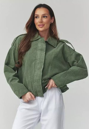 Kobieta o długich brązowych włosach, ubrana w zieloną, oversize'ową kurtkę i białe spodnie, patrząca na bok, z rękami przy talii, na jednolitym tle.