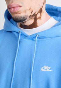 Világoskék, pamutból készült kapucnis pulóver, fehér Nike logóval a mellkasán és ezüst fűzőlyukakkal, húzózsinórral. Puha tapintású és laza dizájnú.