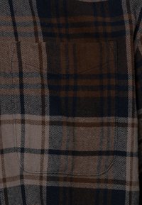 Flanella a quadri, principalmente marrone e blu, presenta una texture spazzolata, una tasca sul petto e linee a contrasto in un motivo a scacchi.