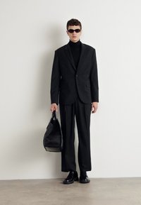 Completo sartoriale nero con vestibilità slim, indossato su un dolcevita nero. Gli accessori includono occhiali da sole neri e una borsa nera con texture. Scarpe nere.