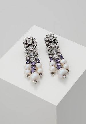 Boucles d'oreilles pendantes longues avec des cristaux clairs, des pierres précieuses violettes et des perles blanches disposées en rangées décroissantes sur une surface blanche inclinée.